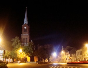 Michał Lutkowski – Czempiń o zmierzchu. Zdjęcie przedstawia fragment rynku w Czempiniu z zabytkową wieżą kościelną kościoła należącego do Parafii Rzymskokatolickiej p.w. Św. Michała Archanioła w Czempiniu. Zdjęcie wykonano podczas spaceru po Czempiniu.